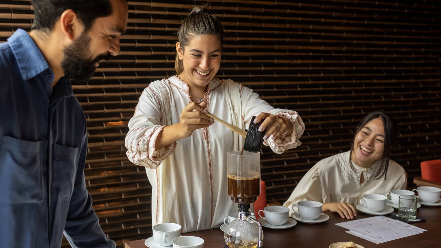 At a table set with white cups and saucers, two smiling guests watch as another pours coffee into a siphon coffee maker.