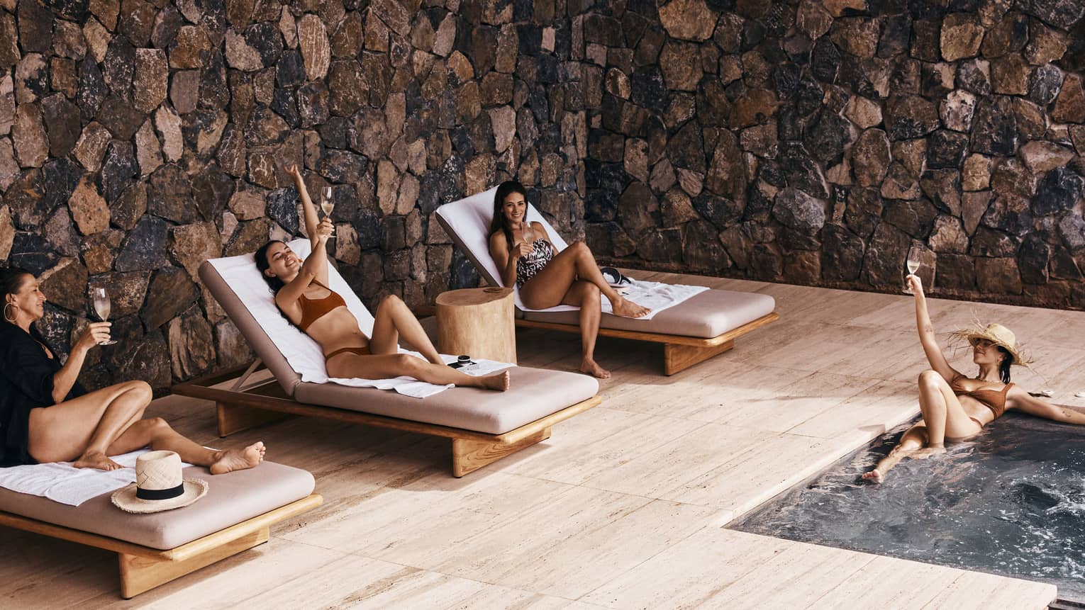 Four elegant women unwind in a soothing pool, surrounded by rustic stone walls, drinking champagne and laughing while creating unforgettable memories