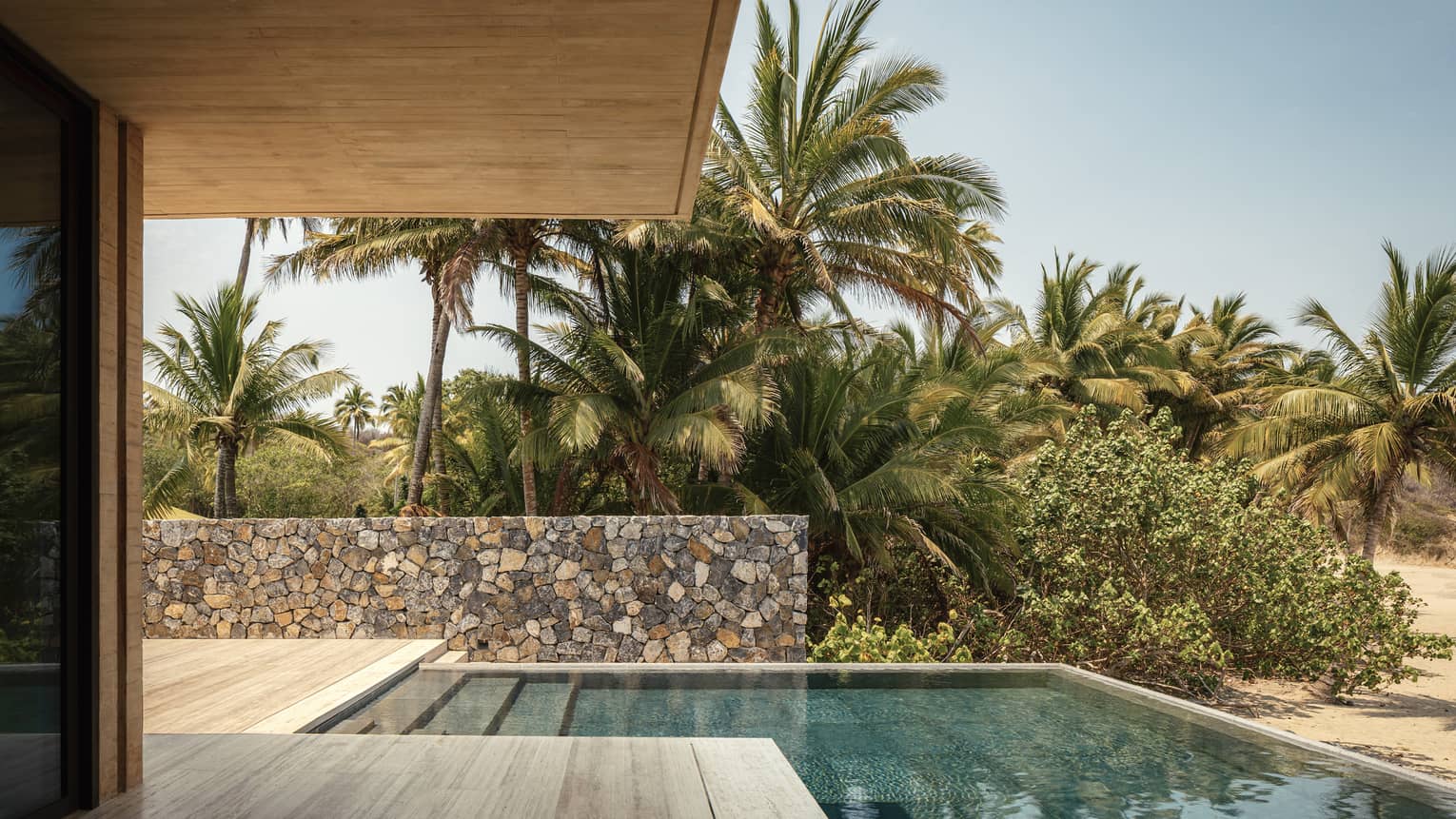 Outdoor poolside area with stone walls, surrounded by lush palm trees and greenery. A covered patio overlooks the tranquil scene under a clear blue sky.