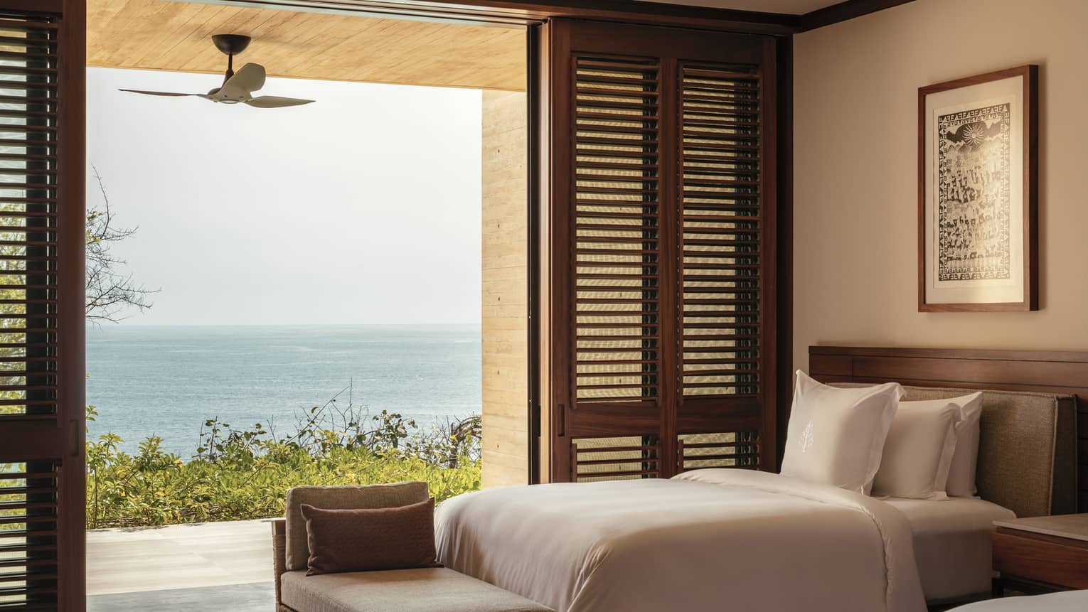 Bedroom with twin beds and white linens, opening to a terrace with ocean views. Wooden shutters and a ceiling fan enhance the room’s airy, tropical feel.