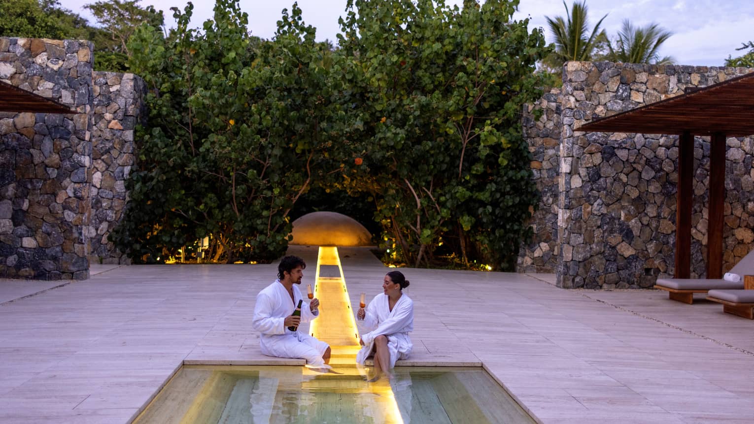 Two guests wearing spa robes and sitting by the spa tub holding wine glasses