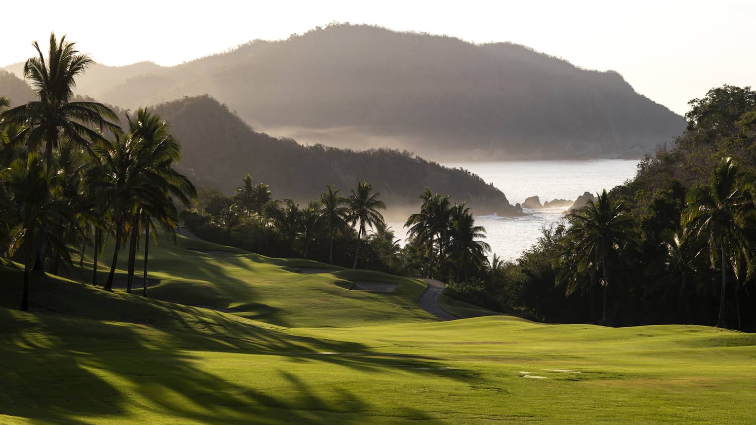Palm trees cast long shadows on a majestic golf course against a backdrop of misty hills jutting out into the bay.