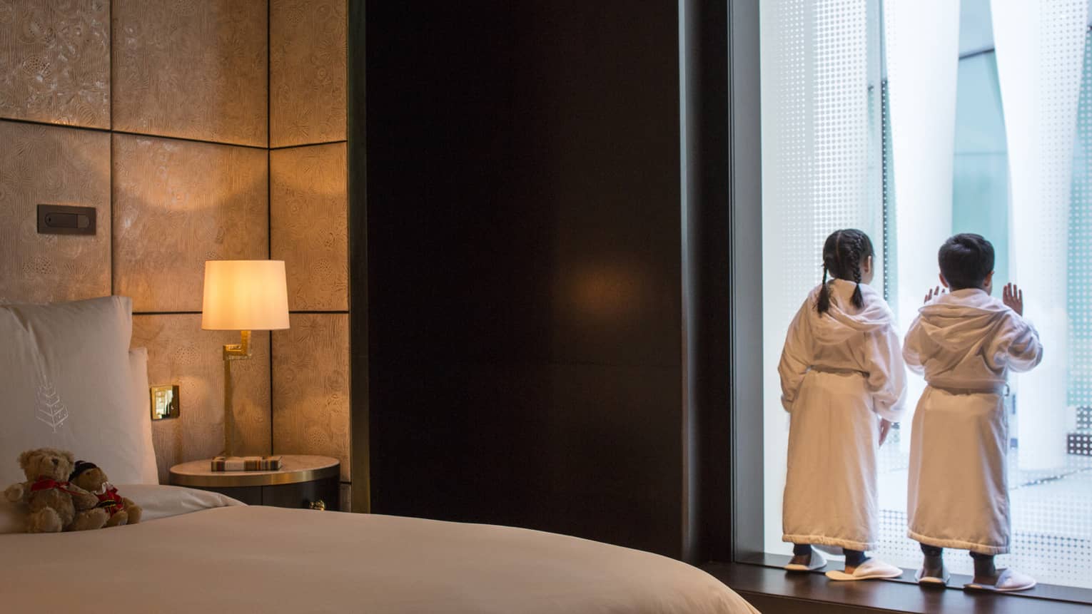 Young boy and girl in bath robes and slippers peer out the window at the city below, two teddy bears on the bed behind them