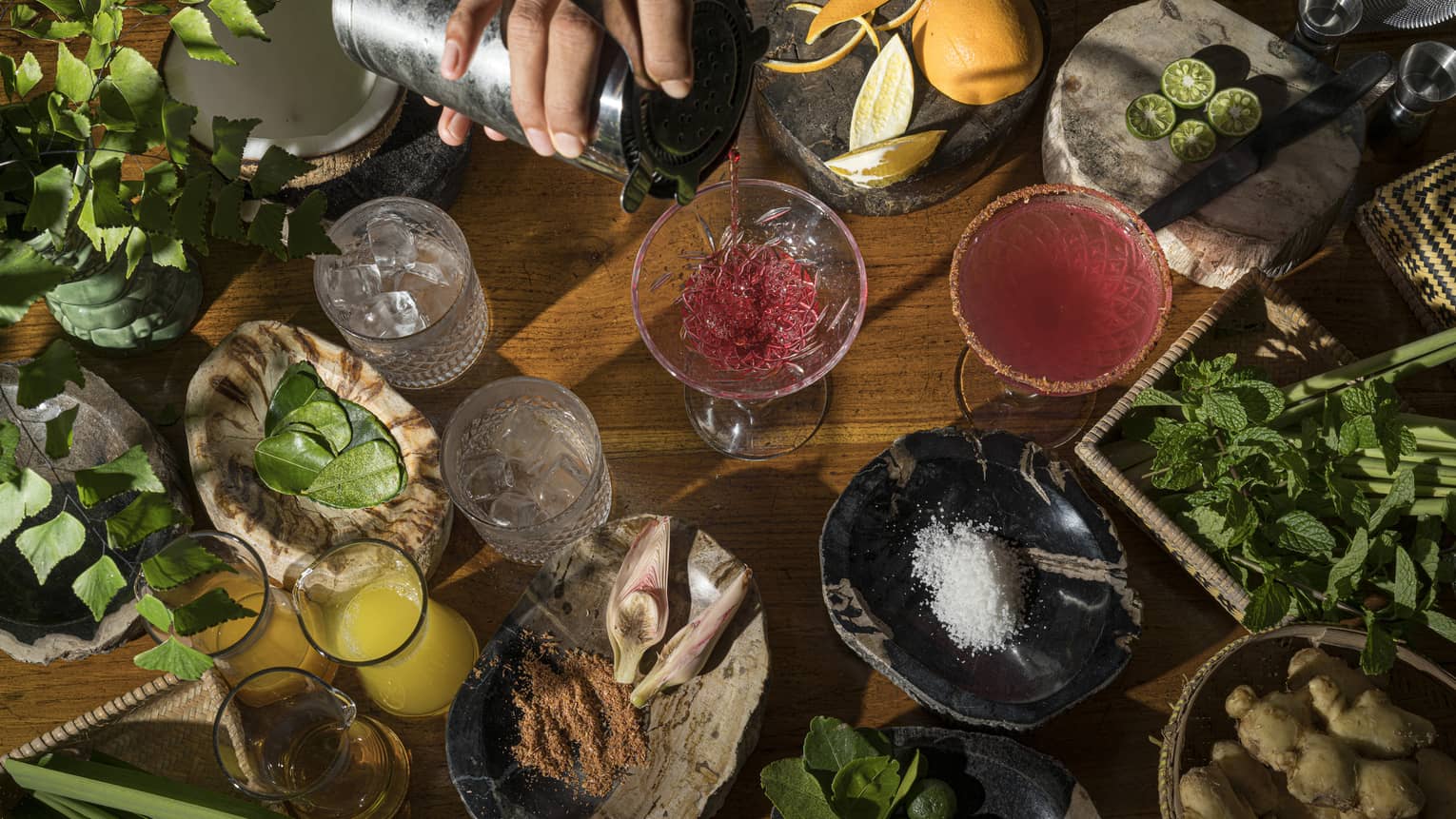 Aerial view of bartender's hand pouring ruby-coloured cocktail intro stemmed glass, local ingredients on wooden bartop surrounding it