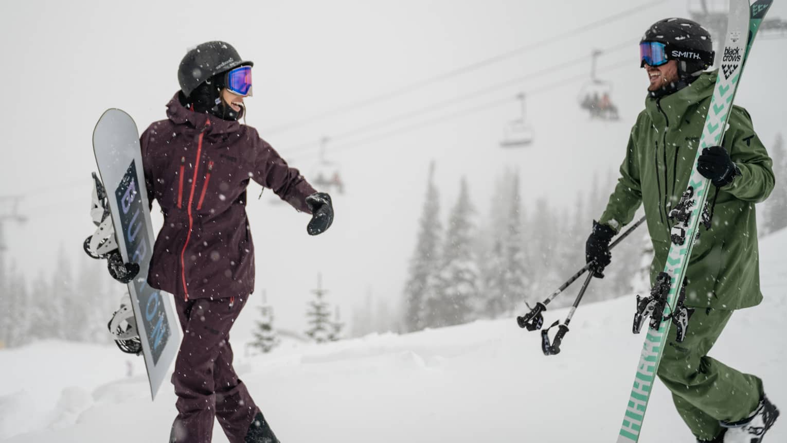 Person wearing burgundy snow suit holding a snowboard walks next to person in a green snow suit holding skis with a snowy landscape and ski lift in the background