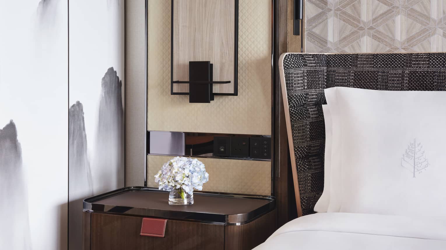 Close-up of a hotel bedside table with a vase of white flowers, next to a bed with white linens. A wall light fixture and geometric accent wall are visible.