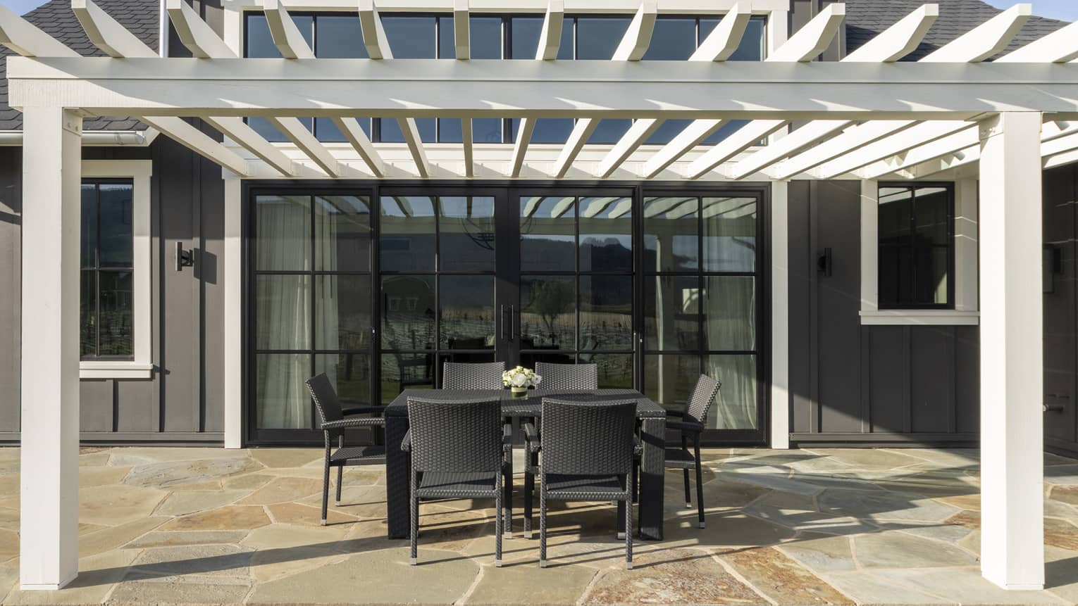 Outdoor villa patio with black wicker dining set under a white pergola, stone flooring, large glass doors and gray siding on the building exterior