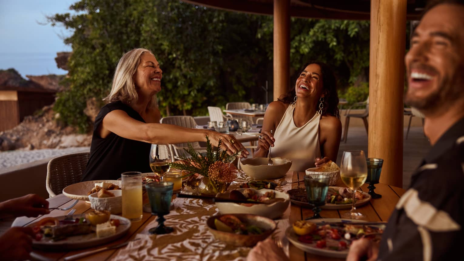 Three people laugh around a dining table on outdoor restaurant terrace