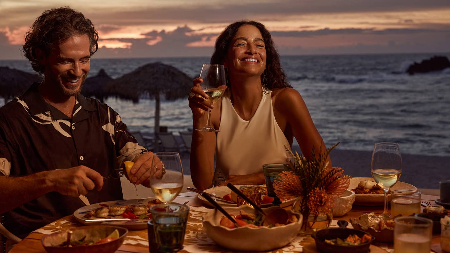 Man and woman laugh while dining at outdoor table filled with food and wine, next to beach and ocean