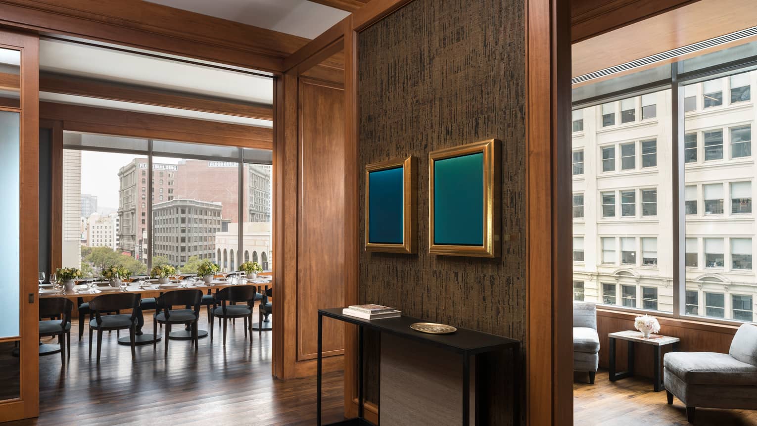 A waiting area in a restaurant with grey chairs and a small table, a private dining area with large windows can be seen through a hall.