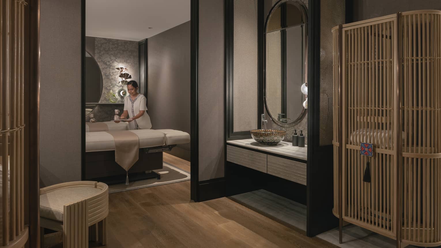 A tray is gently placed on one of two massage tables in a warmly lit spa room with soft grey walls and natural wood décor