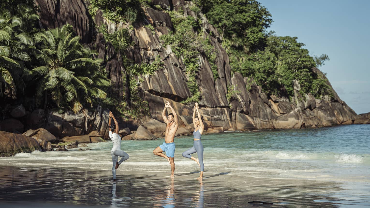An instructor leading a couple through yoga poses on the beach. In the background is a rocky cliff covered in greenery.