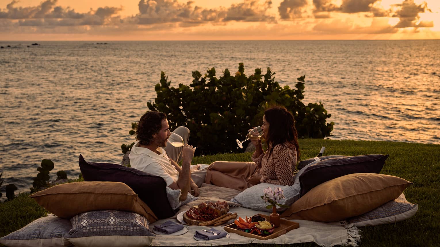A man and woman lay on blanket and pillows during luxury picnic at sunset, next to the ocean