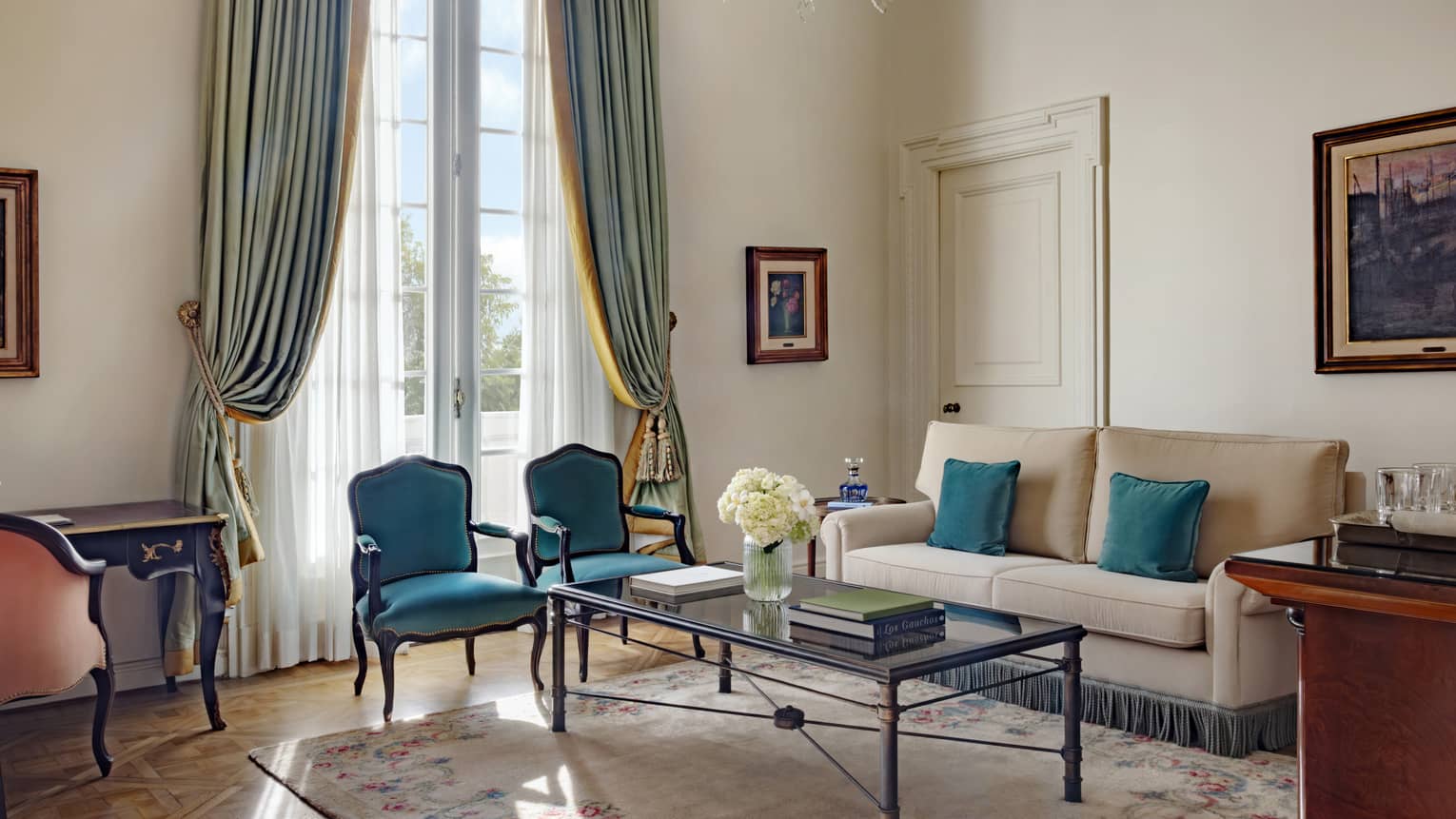 Living room of a hotel suite, featuring a beige sofa, turquoise chairs and draped window curtains curtains