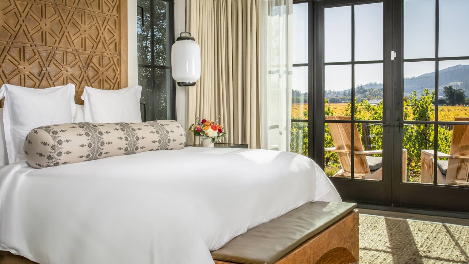 Luxury resort bedroom with a king bed, carved wood headboard, patterned bolster pillow, pendant light, bench and doors opening to a vineyard-view terrace