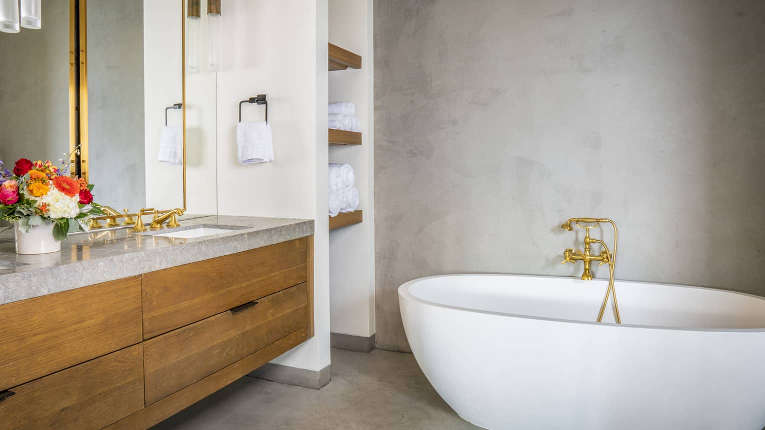 Luxury resort bathroom with a freestanding white bathtub, wooden vanity with gray countertop, floral arrangement, golden fixtures and built-in towel shelves