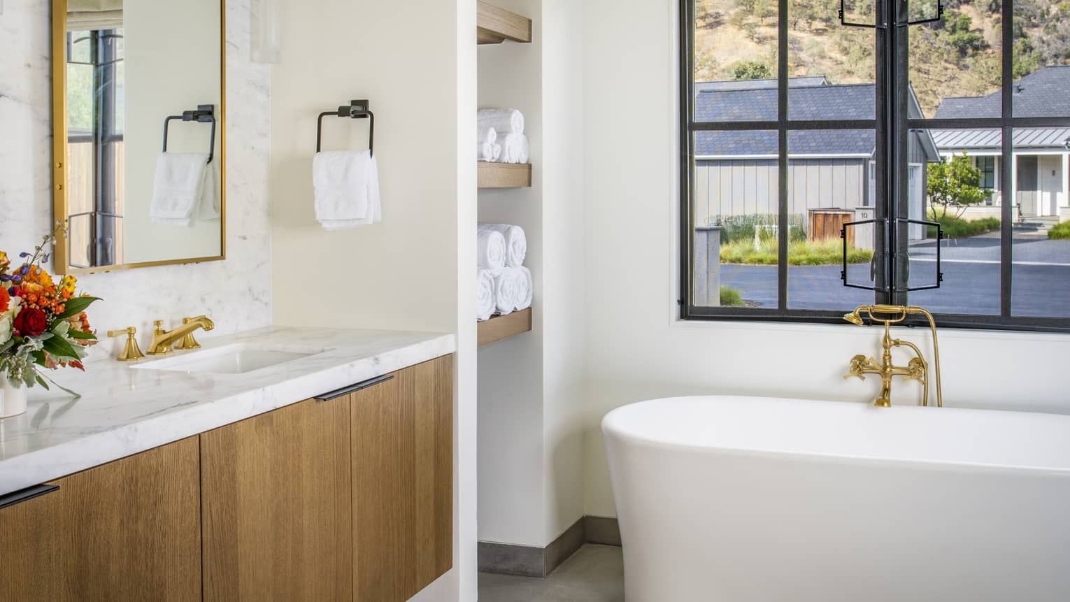 Bathroom with freestanding bathtub, marble countertop vanity, gold fixtures, floral arrangement, built-in towel shelves and window overlooking the outdoor area