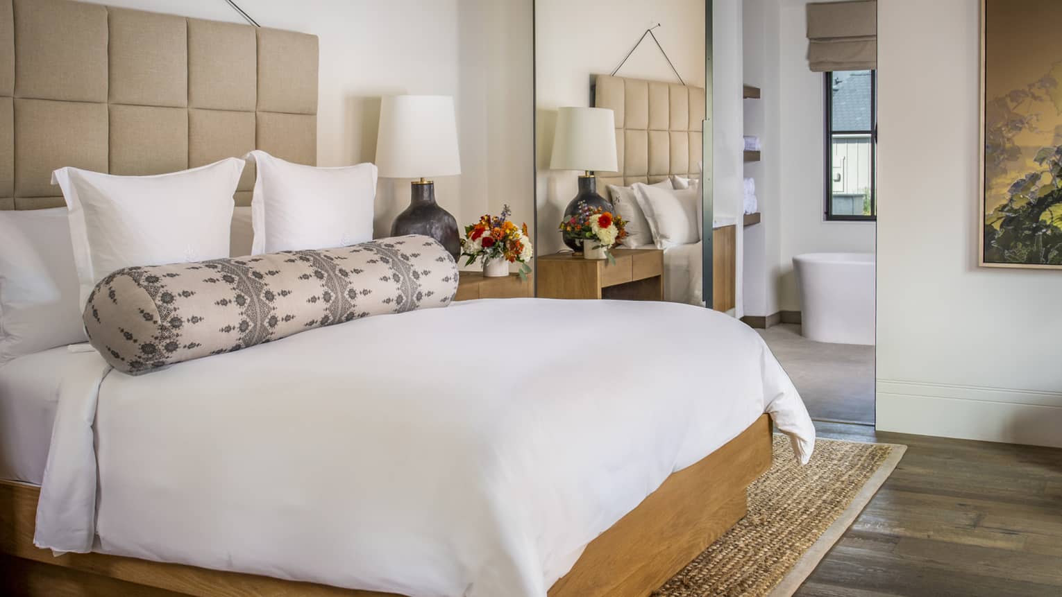 Resort bedroom with a king bed, beige upholstered headboard, patterned bolster pillow, wood nightstands with lamp and flowers and a view into bathroom with tub