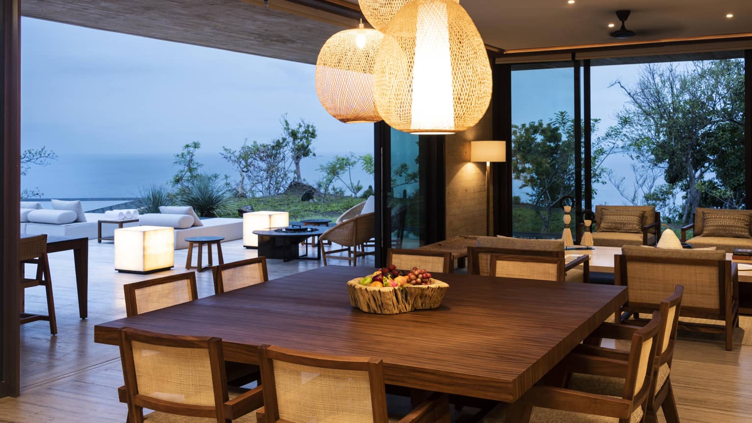 Modern dining room with a large wooden table, wicker chairs and woven pendant lights. Sliding glass doors open to an outdoor seating area with ocean and garden views.