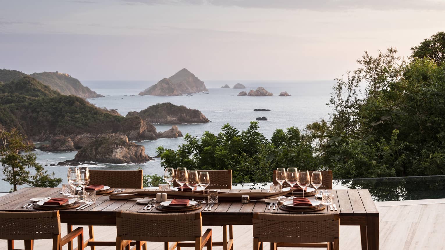 A beautifully set outdoor dining table with glassware and plates, overlooking a breathtaking ocean view with rocky islands and lush greenery at sunset