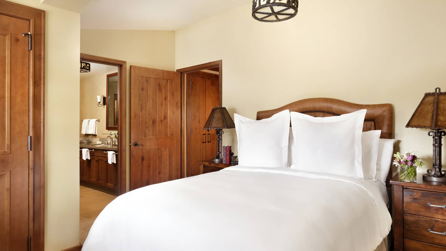 Bedroom with a leather headboard, dark wood side tables with rustic lamps and floral decor, and a view into a connected bathroom