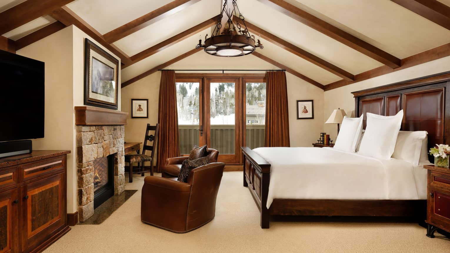 Bedroom with a vaulted wood-beam ceiling, stone fireplace, king bed, leather armchairs and a TV stand, opening to a balcony with snowy scenery view