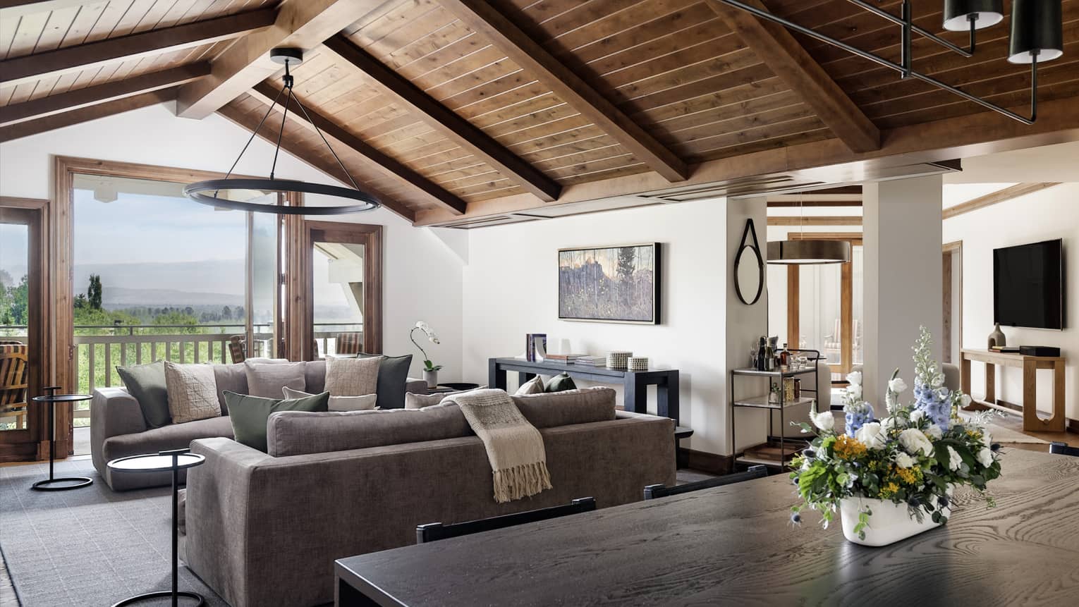 Living and dining area with vaulted wood ceiling, two grey sofas, dining table with a floral centrepiece, and mountain views