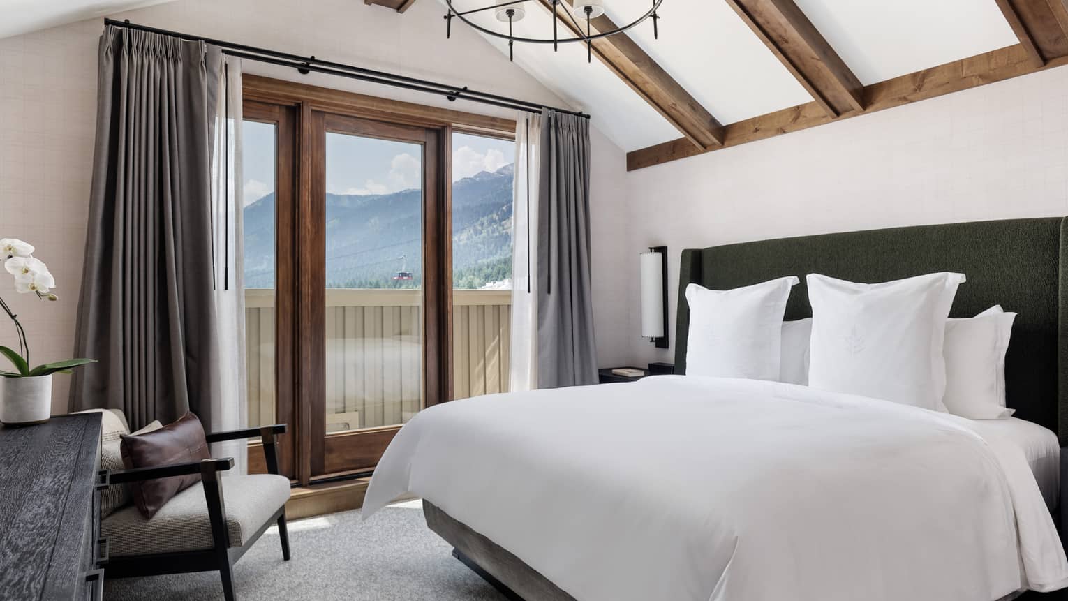 Luxury resort bedroom with king bed, arm chair, vaulted ceiling with wooden beams, and walk-out balcony with mountain view