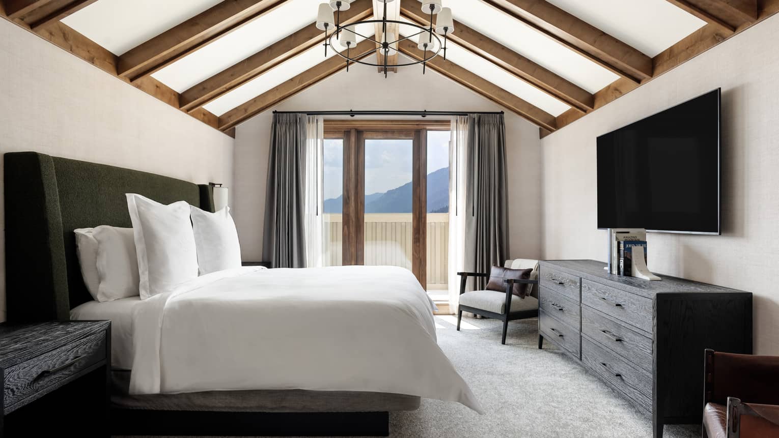 Luxury resort bedroom with vaulted wood-beam ceiling, king bed, dresser, TV, armchair and balcony with mountain view
