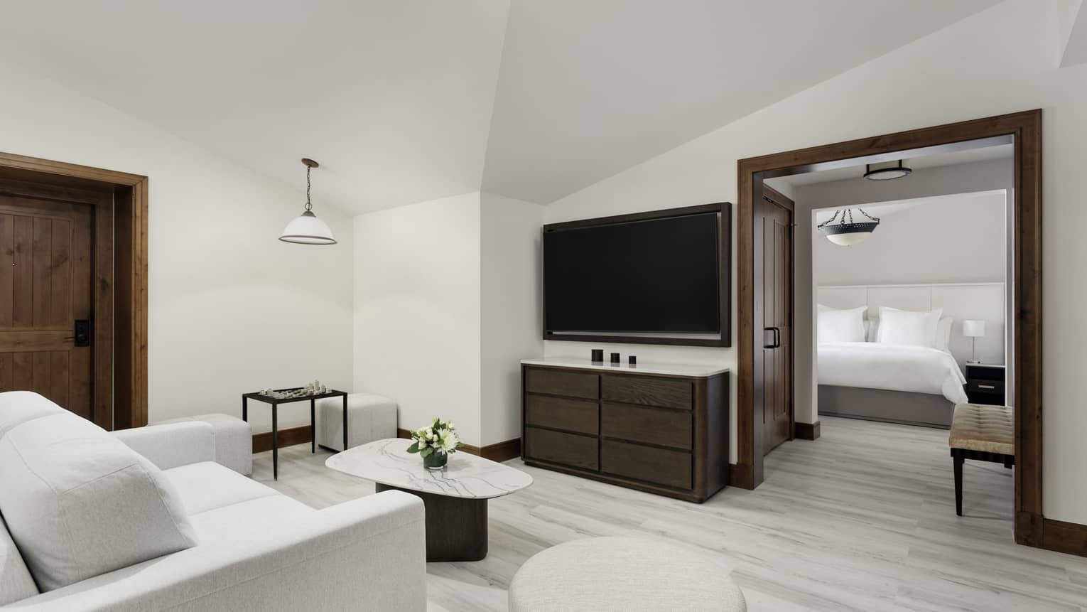 Small living room with a TV, dresser, marble coffee table and grey sofa, leading into a bedroom with king bed