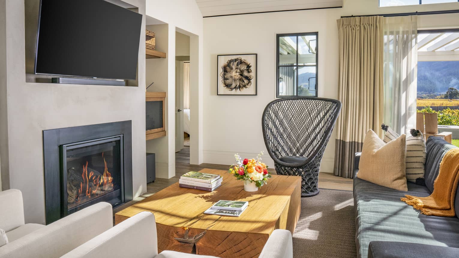 Bright living room with a fireplace, wall-mounted TV, wooden coffee table, black wicker chair, gray sofa, beige armchairs and views of vineyards and mountains