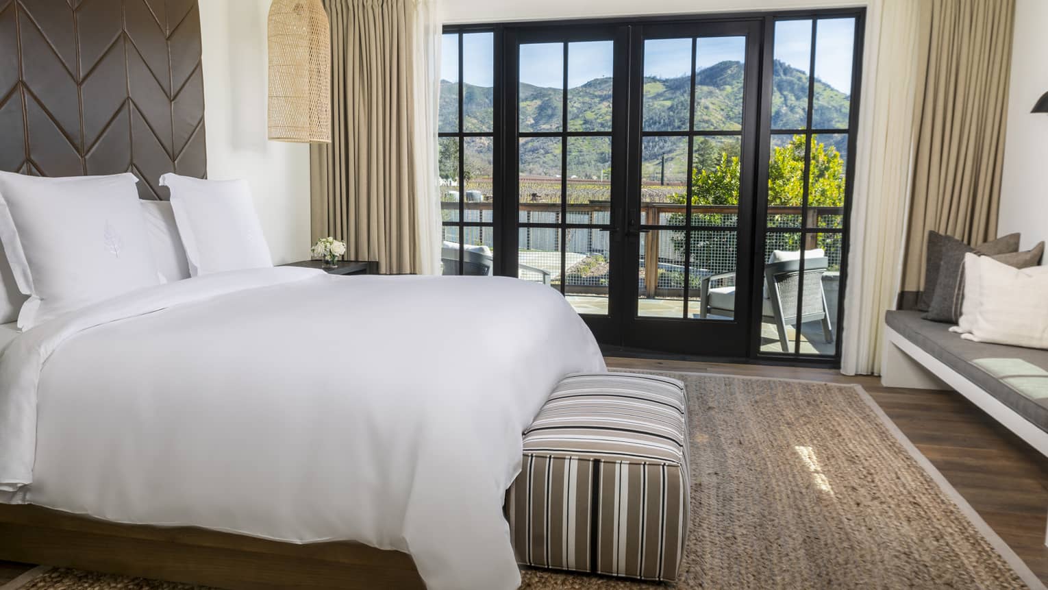 Luxury resort bedroom with a king bed, dark textured headboard, striped bench, built-in seating and large glass doors opening to a mountain-view terrace