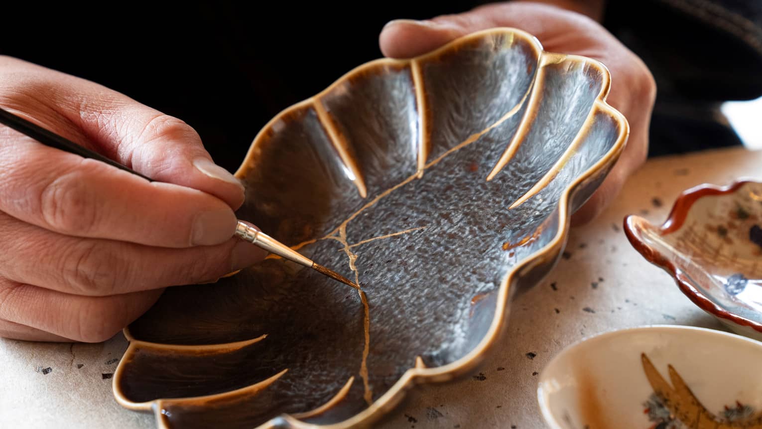 A person using the art of Kintsugi, by using gold paint to paint over the cracks of a once broken black flower shaped plate.