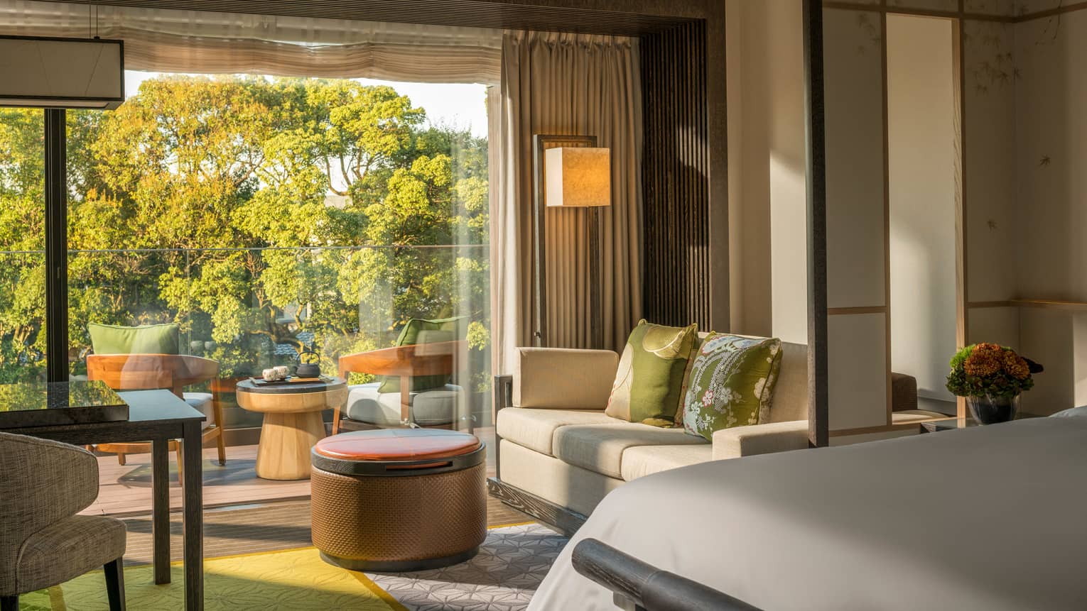 Guest room with king bed, sofa, desk and chair, round ottoman and private balcony with a seating area overlooking a pond garden