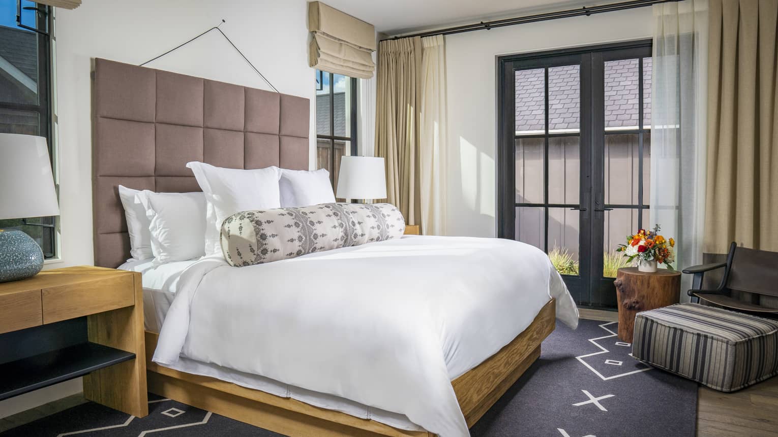 Resort bedroom with a king bed, upholstered headboard, patterned bolster pillow, wooden nightstands, chair with striped ottoman and French doors with curtains