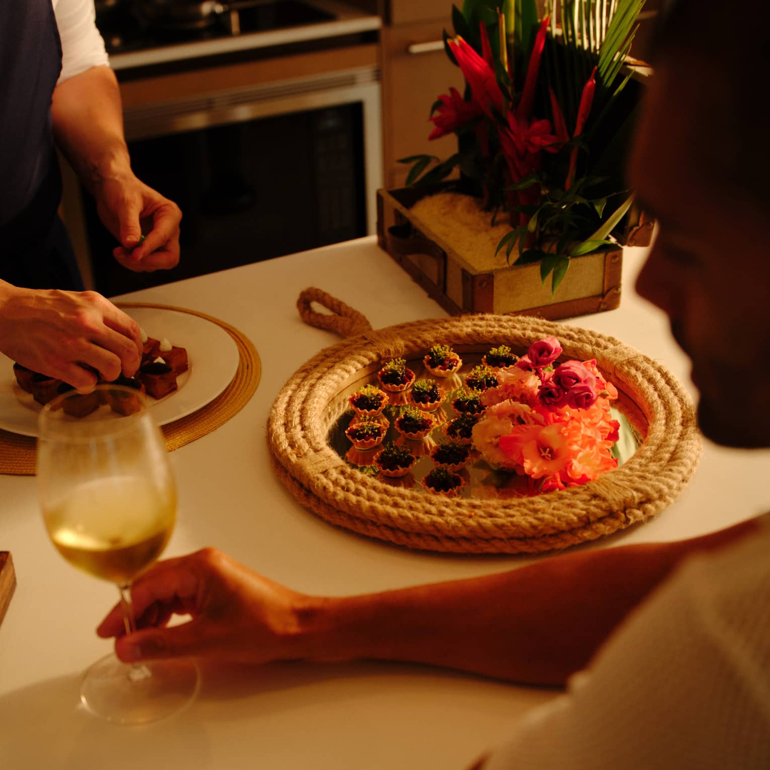 Person sits at a kitchen counter holding a glass of white wine as a chef prepares a plate of appetizers,Person sits at a kitchen counter holding a glass of white wine as a chef prepares a plate of appetizers