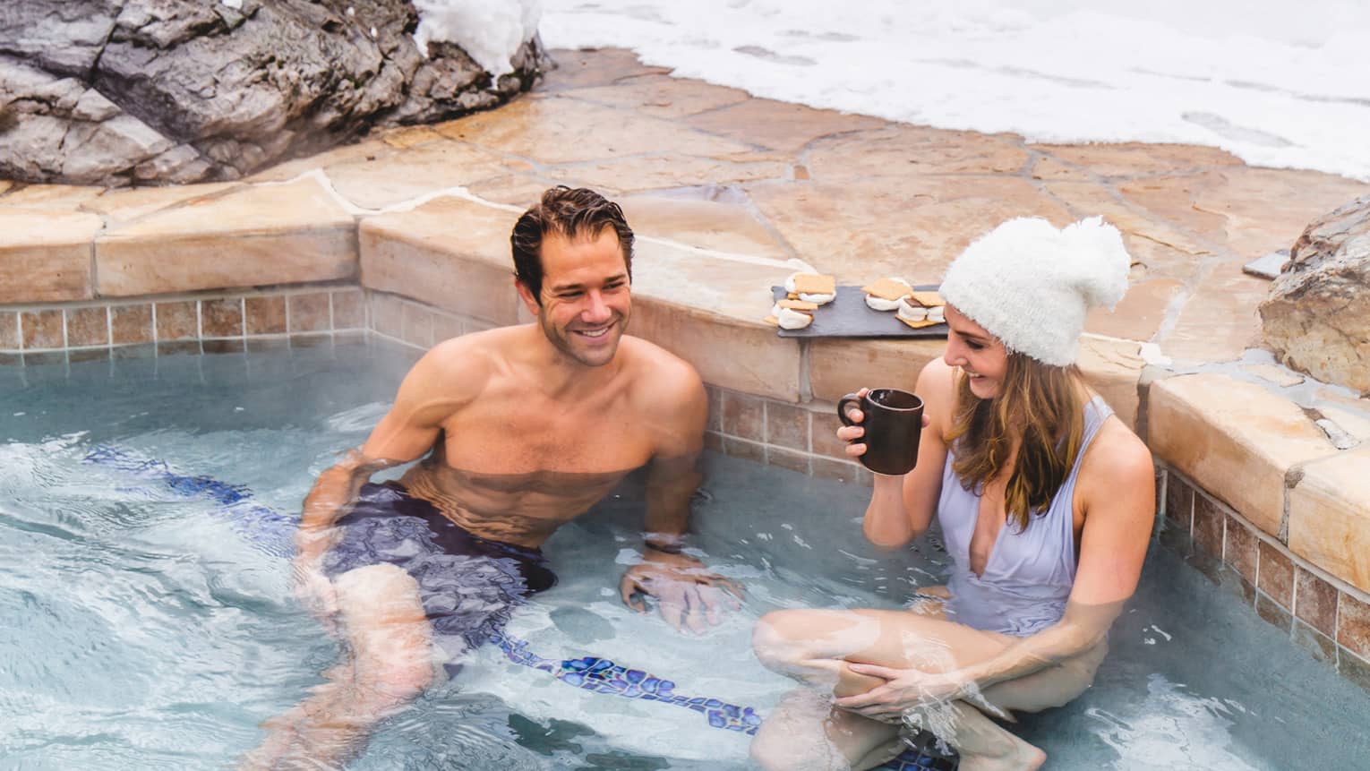 A man and woman in lounging in a heated pool surrounded by snow.