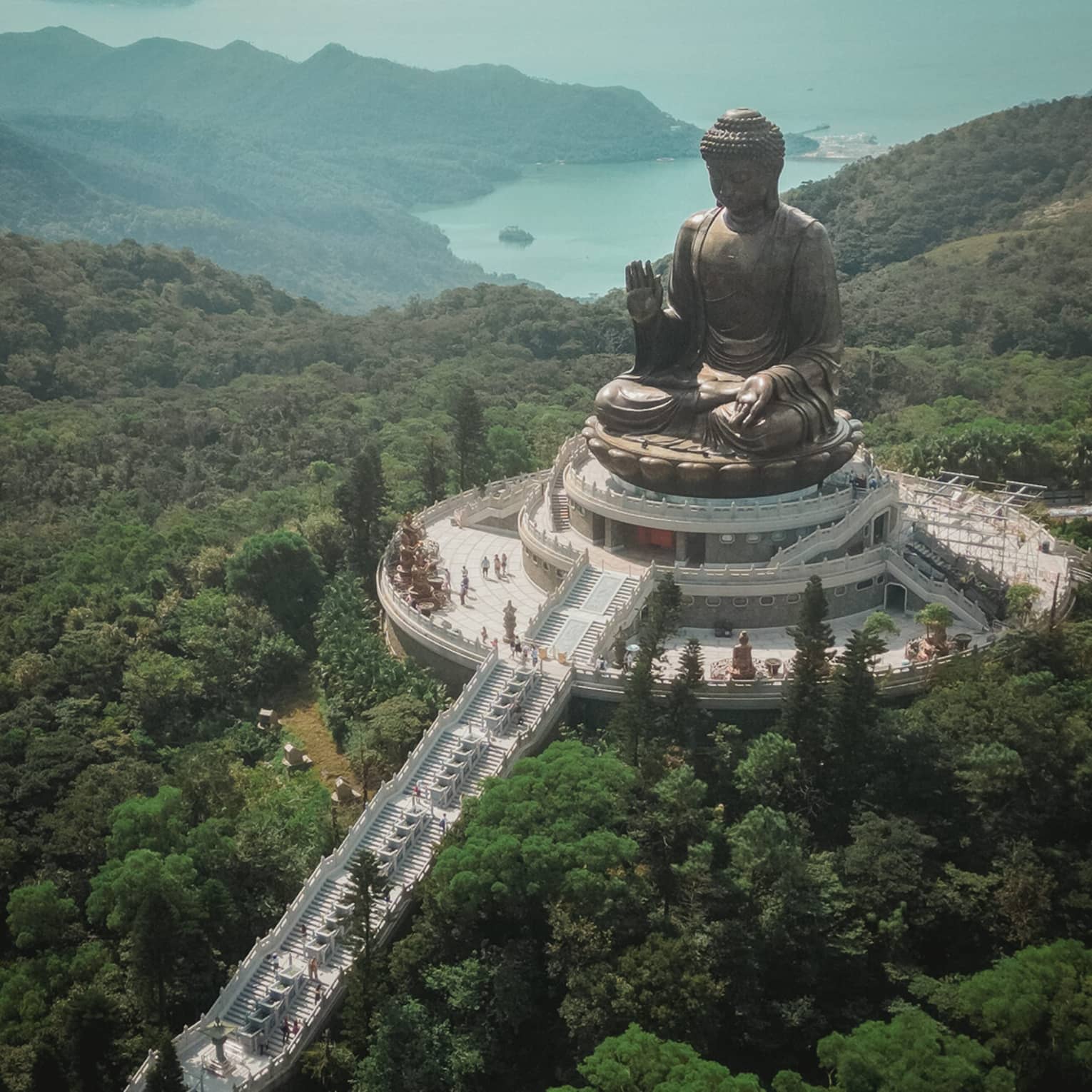 Aerial view of a giant bronze Buddha statue at Po Lin Monastery, surrounded by greenery and mountains. A long staircase leads to the statue on a platform