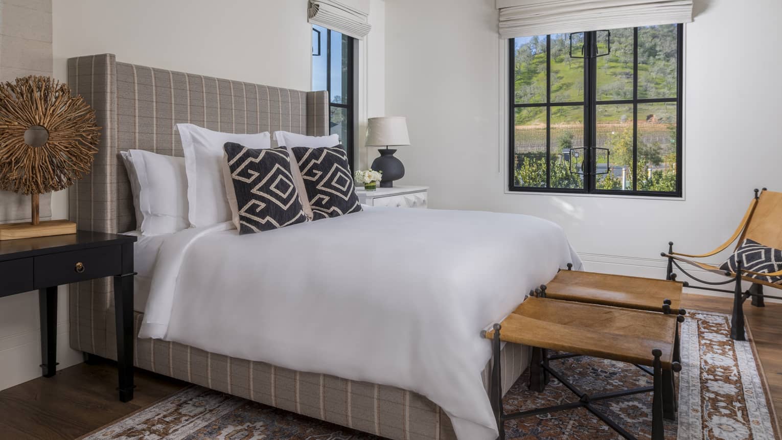 Resort bedroom with plaid upholstered bed, patterned pillows, nightstands, two wooden tables and leather sling chair by the window with hillside view