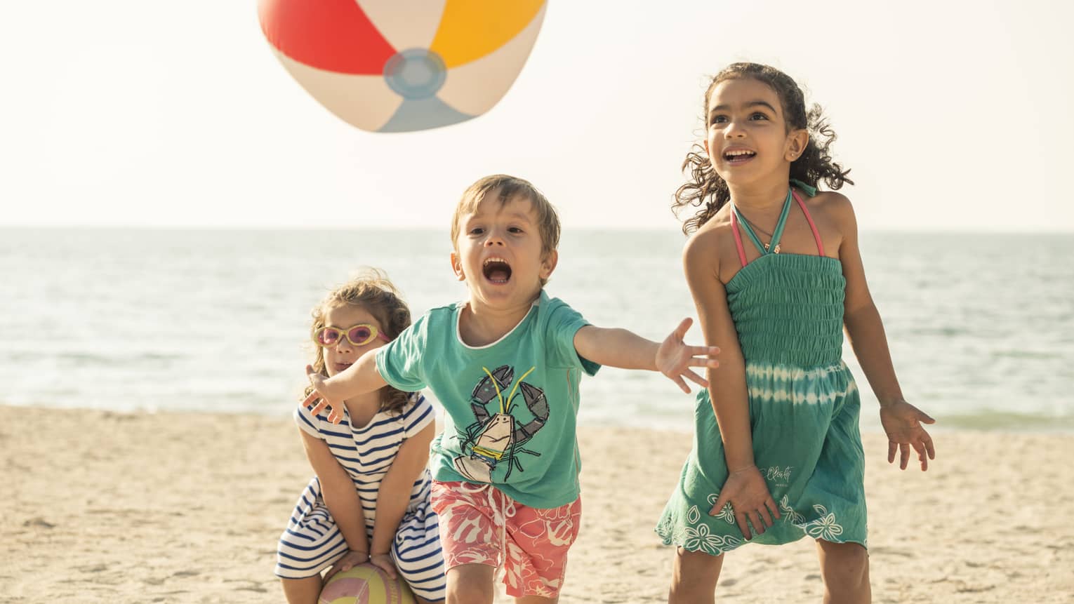 Close-up of three children playing with a beach ball on a sandy beach, the calm ocean and clear sky spanning behind them.