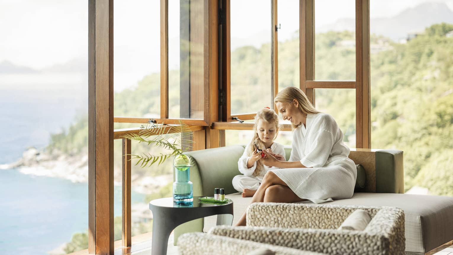 A parent and child wearing white spa robes, sitting on lounge chair with mountain and water views in backdrop