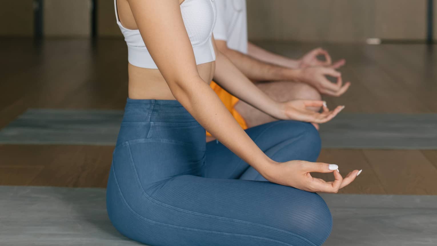 Woman with dark hair tied up in a ponytail and wearing a white tank top and blue yoga pants sits cross-legged on a yoga mat with her hands on her knees and eyes closed, a man wearing a white t-shirt and orange shorts does the same behind her