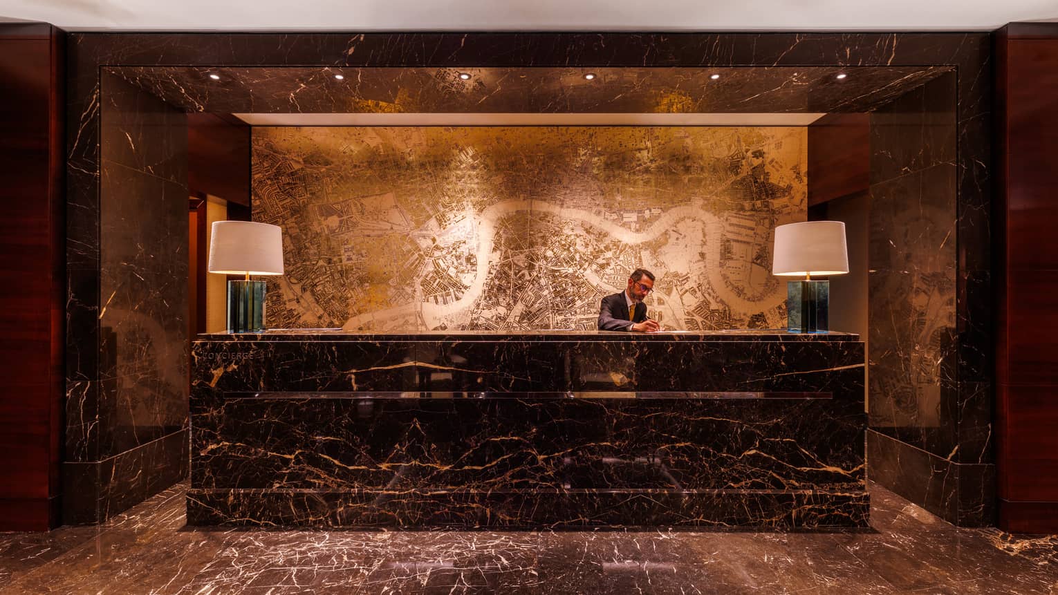 A warm, softly lit hotel lobby space with a person behind a large desk, wall decor, and both desktop and ceiling lighting