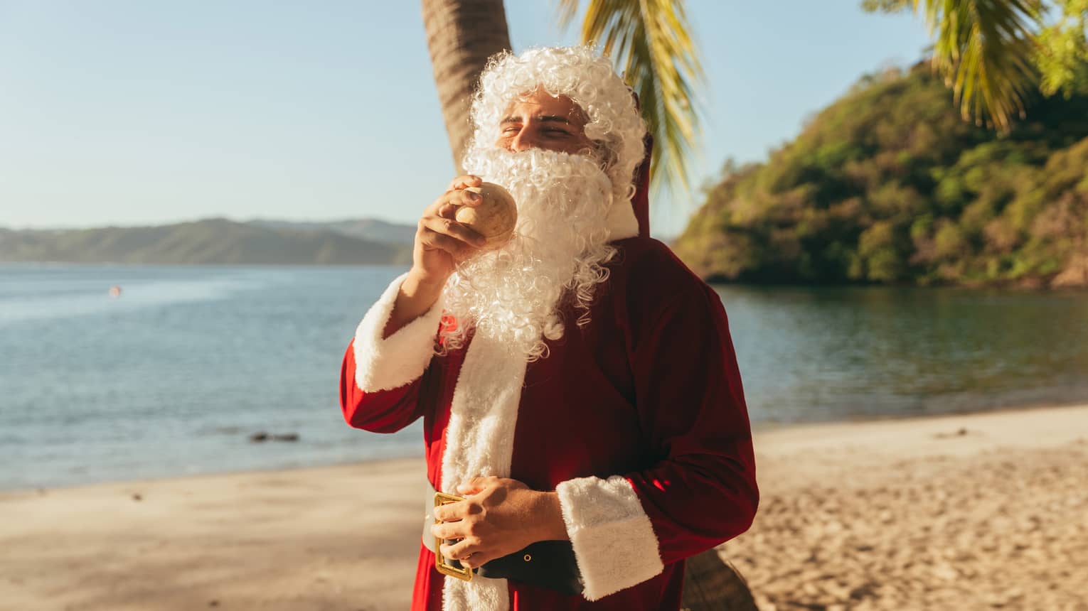 A person on the beach wearing a Santa Claus costume and holding a coconut