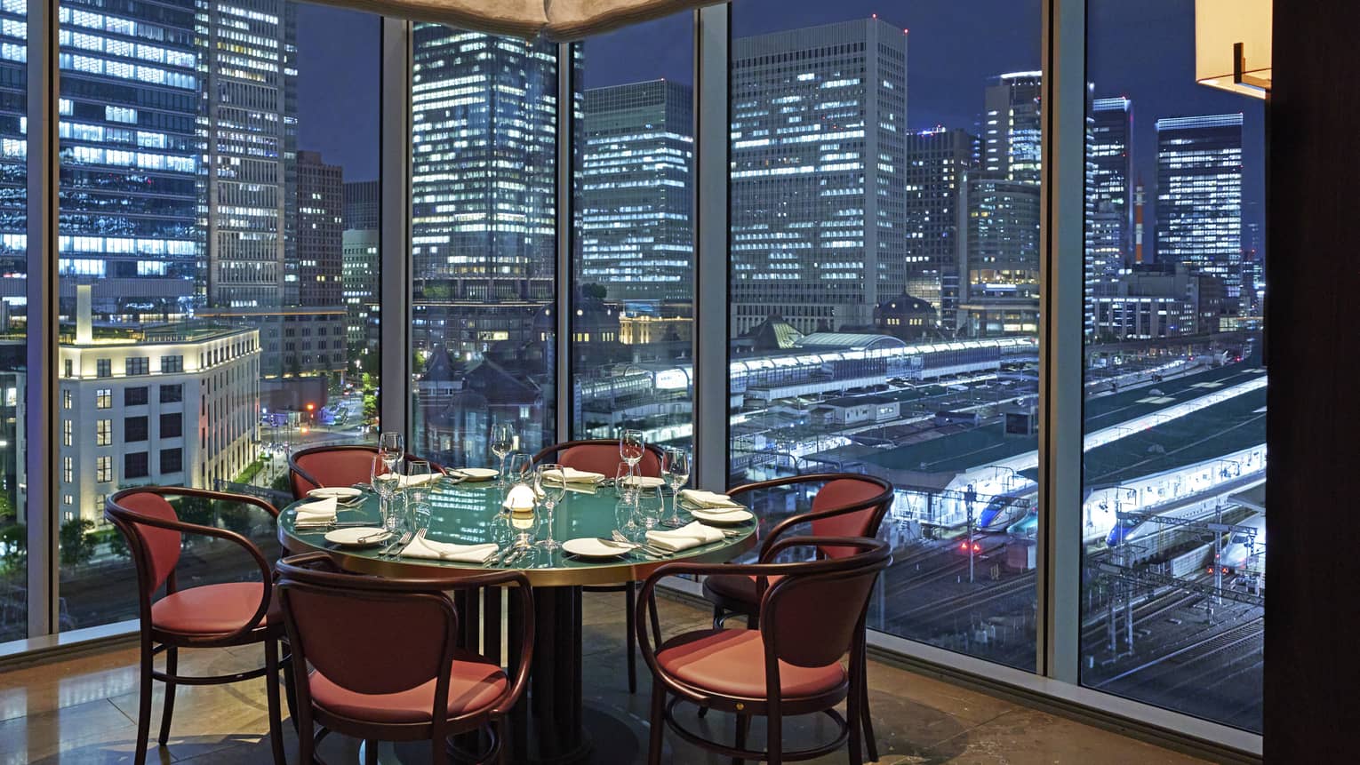 Luxury boutique hotel dining setup with a round table elegantly set for guests, offering a panoramic city view at night through tall glass windows