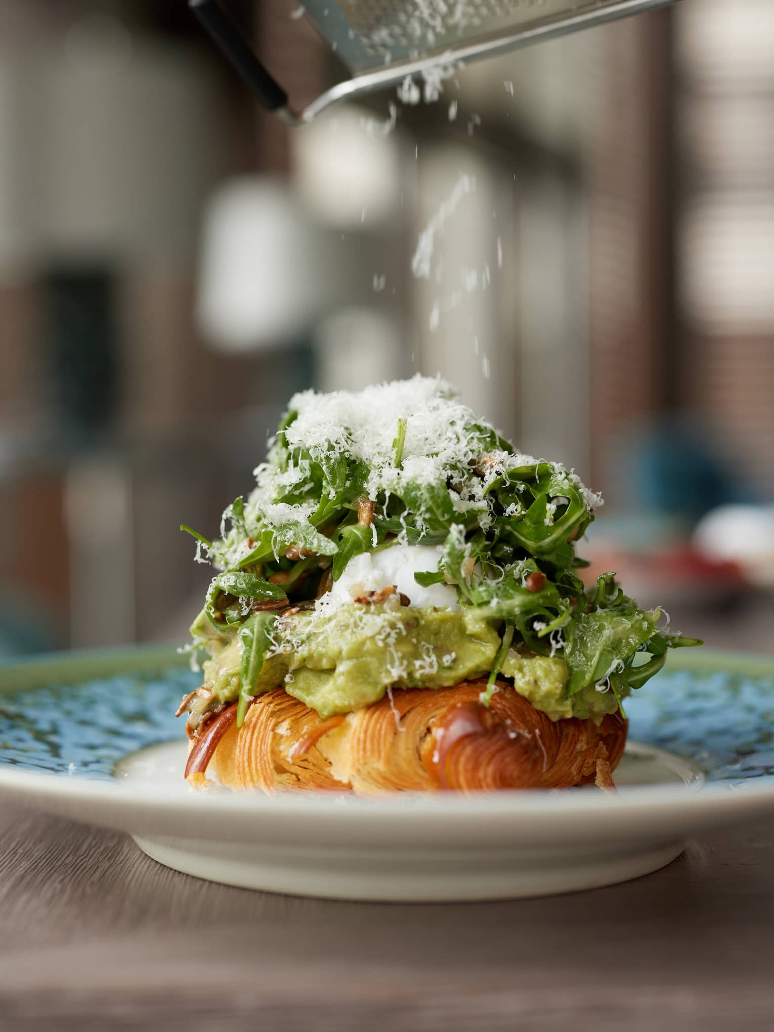 A close-up of a plate holding a croissant covered with avocado, egg and arugula salad topped with grated parmesan.