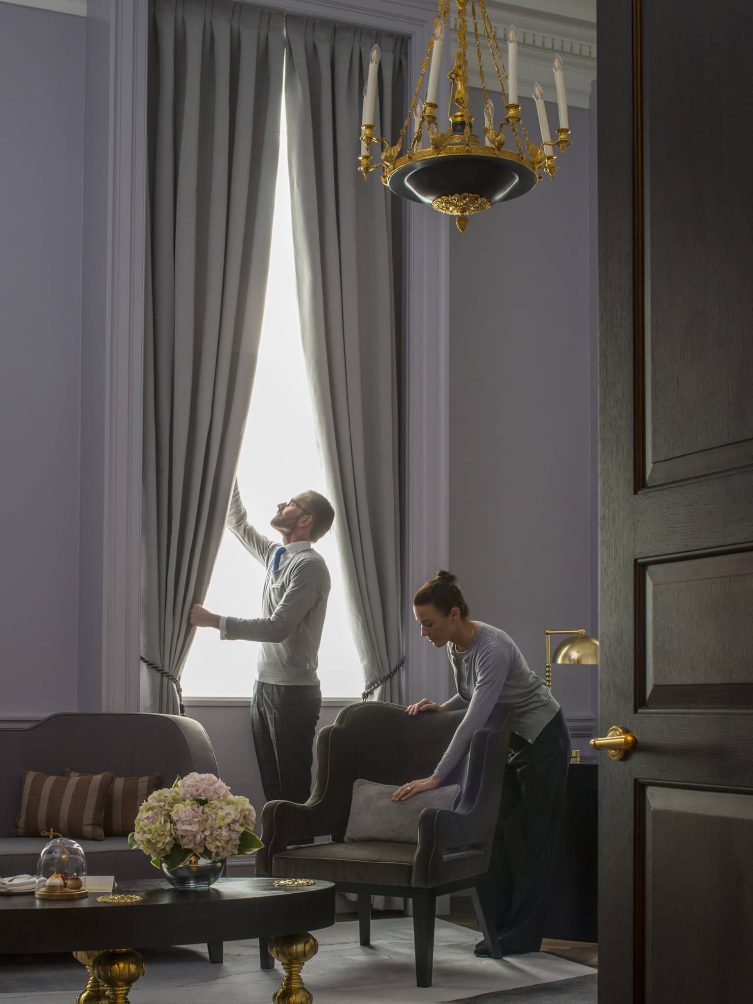 Grand Heritage Suite, man opens drapes as woman smooths pillow on armchair under chandelier