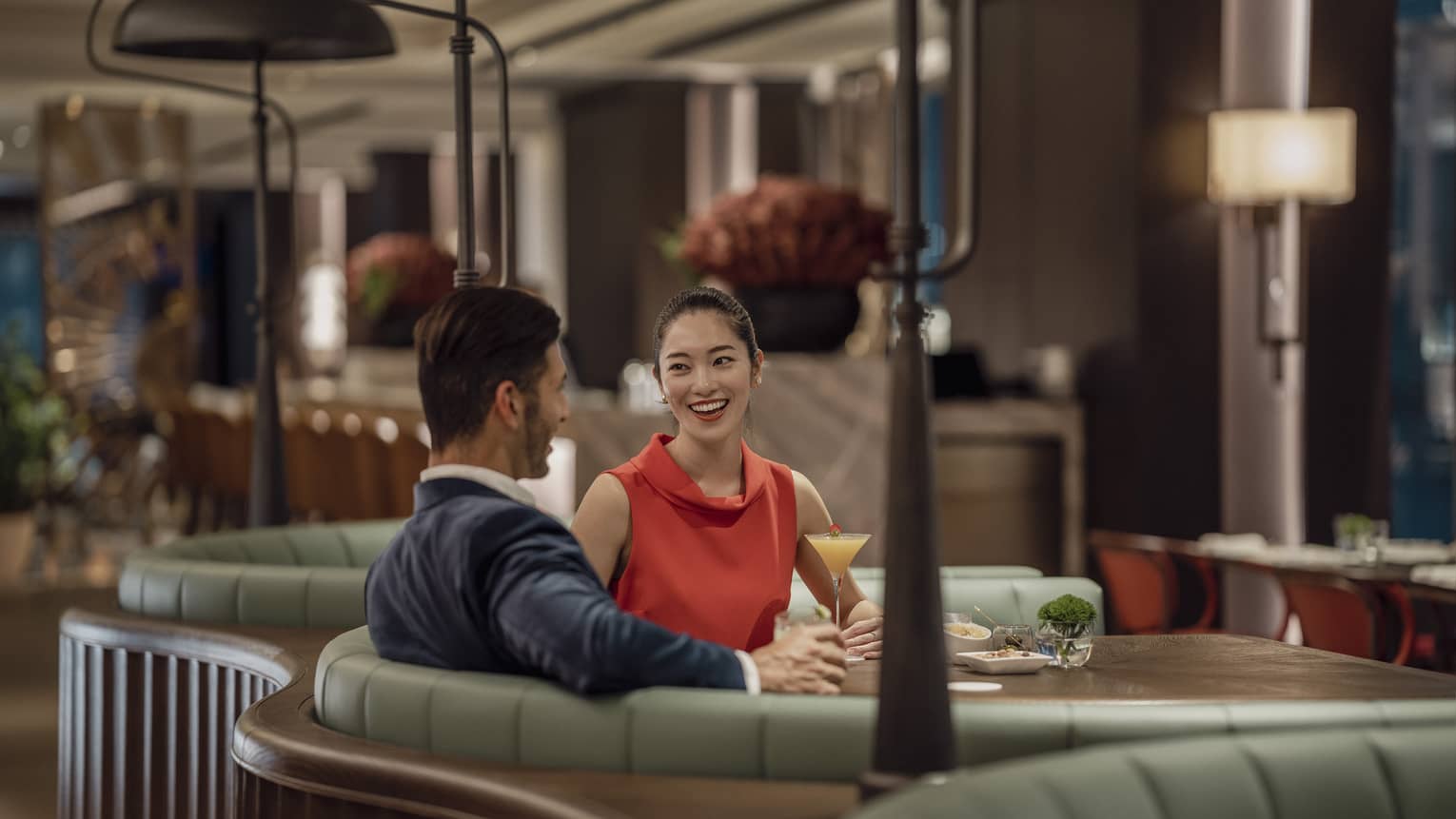 A smiling couple sit at a circular booth in an empty, dimly lit restaurant with cocktails and appetizers on their table.