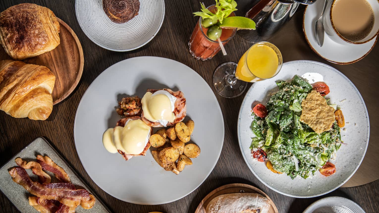 An elaborate brunch, including eggs Benedict, potatoes, bacon, various pastries, salad, a bloody mary and mimosas