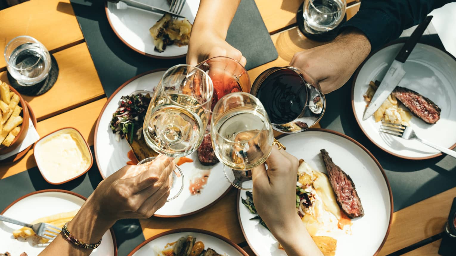 Close-up from above of four hands clinking glasses at the centre of a spread of plates of rare grilled meat and vegetables.